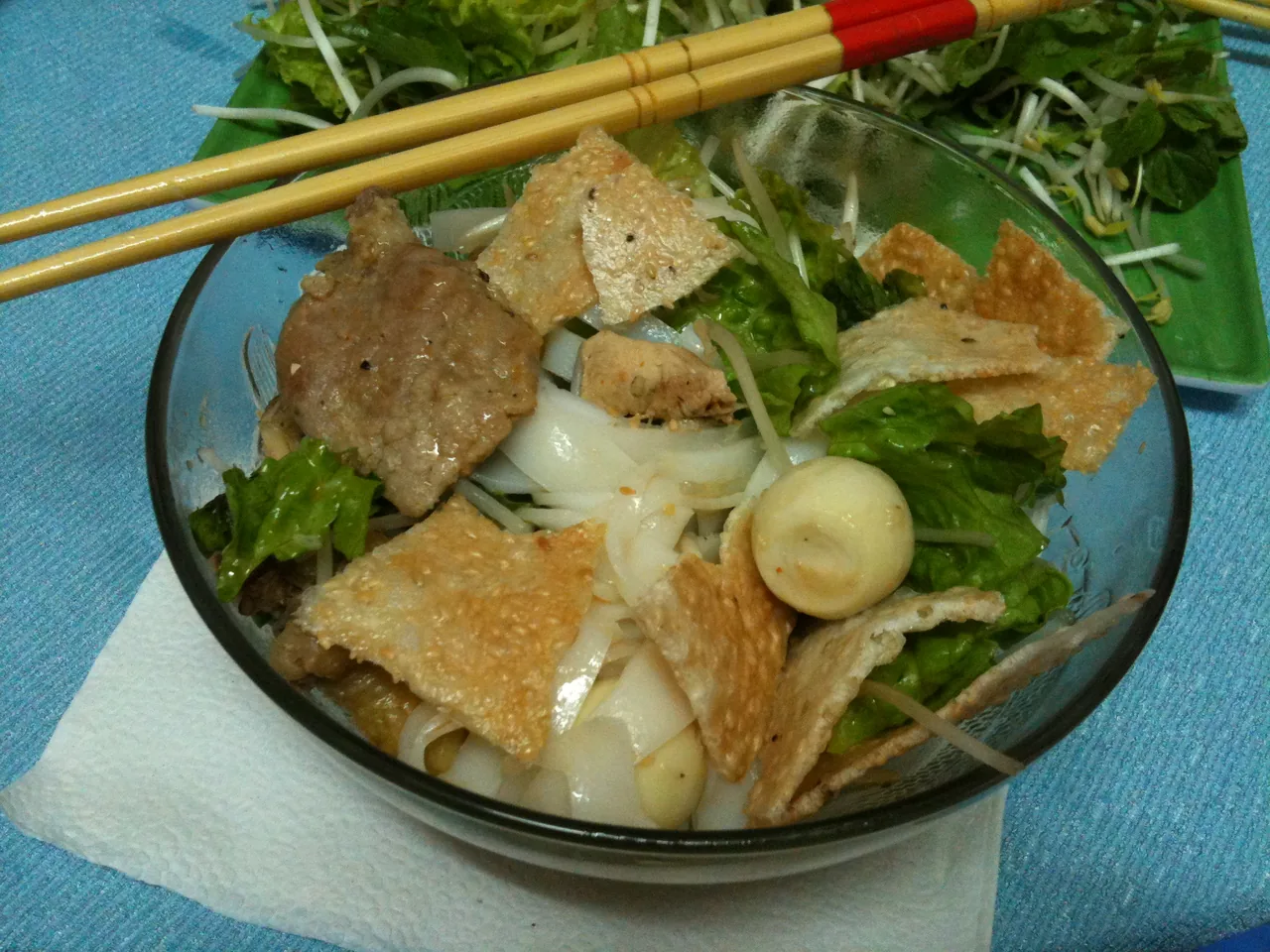 A bowl of Mì Quảng showing wide flat noodles, herbs and a sesame rice cracker
