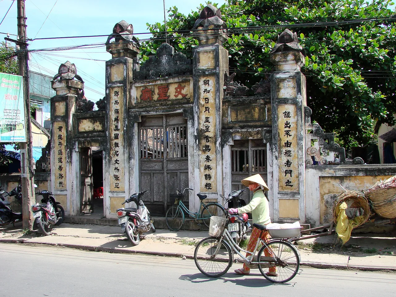 Một phụ nữ dắt xe đạp đi qua những chiếc xe máy đậu bên cổng cổ ở Hội An Một phụ nữ dắt xe đạp đi qua những chiếc xe máy đậu bên cổng cổ ở Hội An