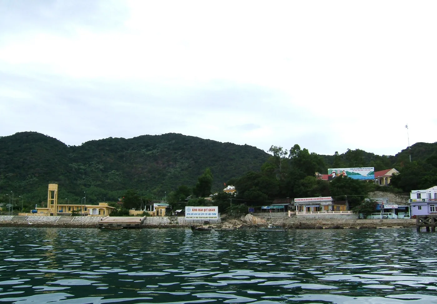The village of Cù Lao Chàm seen from the water, with a pier and a red and white welcome sign on the waterfront