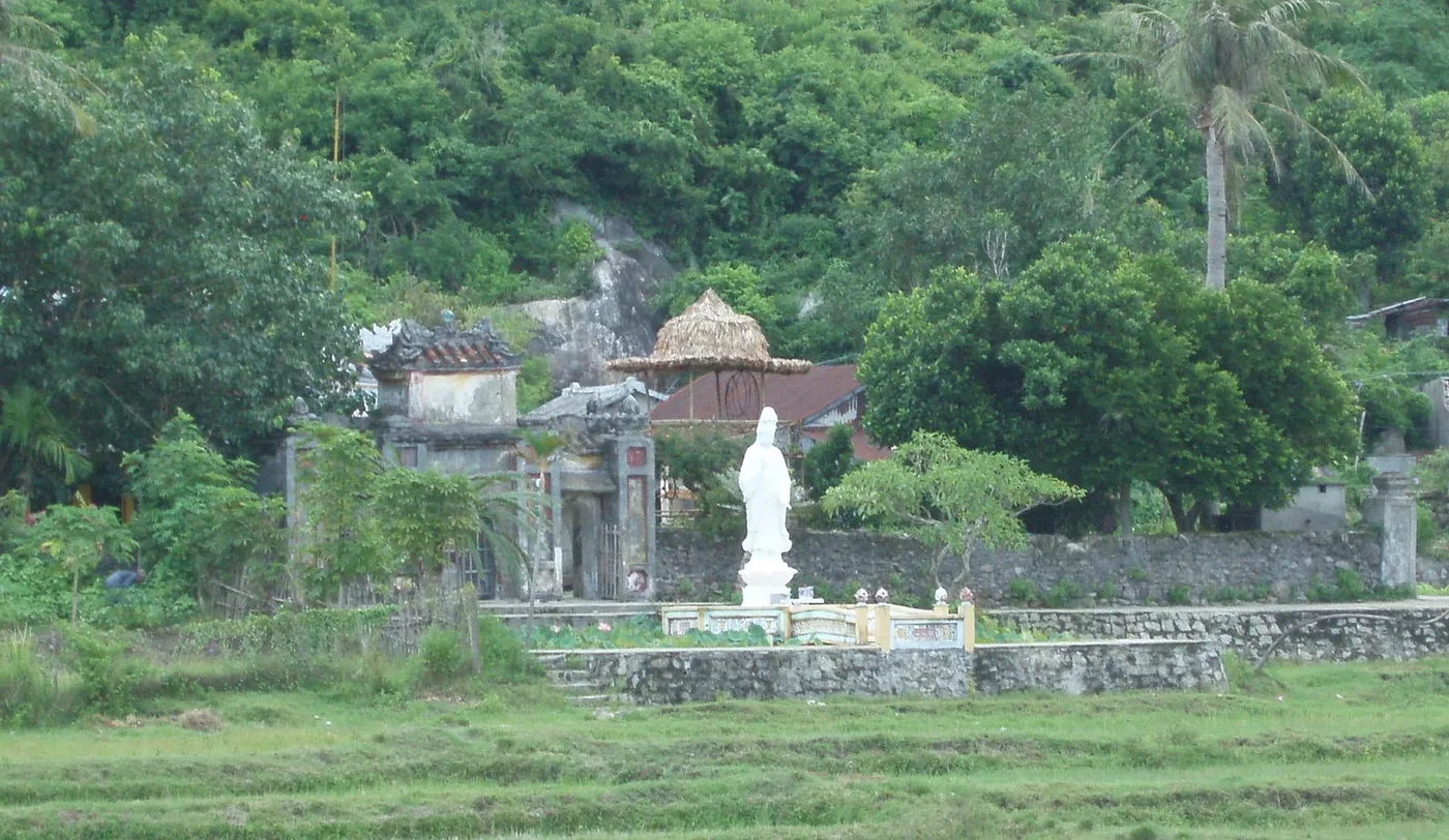 An old weathered temple gateway and a white standing Buddha statue on a plinth seen across a grassy field