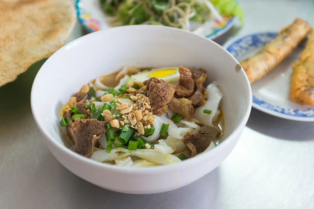 A bowl of mi Quang - flat turmeric-yellow noodles, herbs, peanuts and a sesame rice cracker - on a steel table A bowl of mi Quang - flat turmeric-yellow noodles, herbs, peanuts and a sesame rice cracker - on a steel table