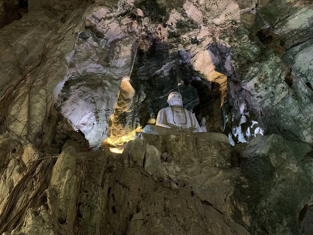 A seated Buddha inside Huyen Khong cave at the Marble Mountains, with uplit marble walls and roots hanging through the opening in the ceiling A seated Buddha inside Huyen Khong cave at the Marble Mountains, with uplit marble walls and roots hanging through the opening in the ceiling