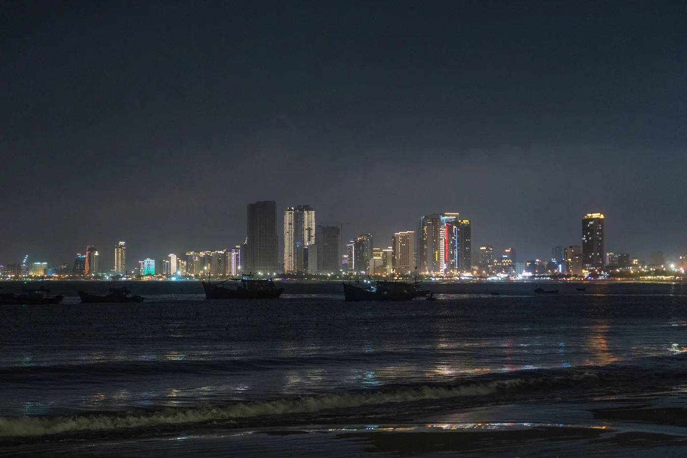 A Da Nang shoreline at night with breaking waves and wet sand in the foreground, moored wooden fishing boats mid-frame and the lit city skyline on the horizon