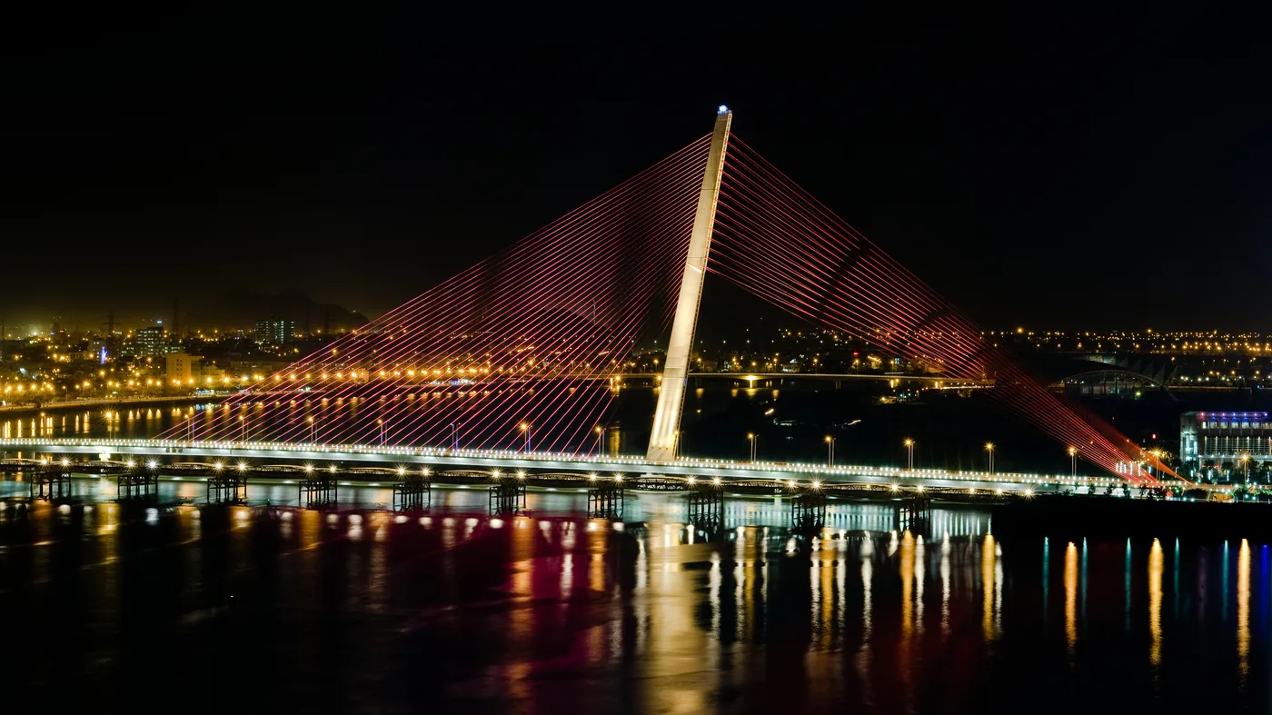Tran Thi Ly Bridge in Da Nang at night from above, lit red and pink, showing its single leaning pylon and fan of cables