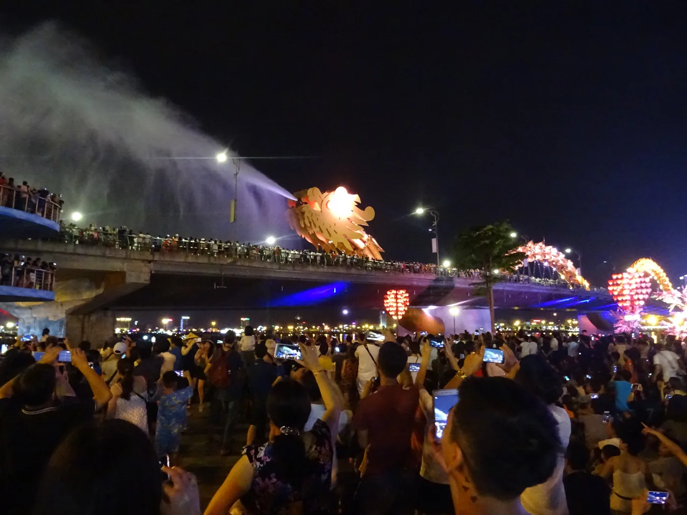 The Dragon Bridge at night with the dragon head spraying a white plume of water over a crowd of hundreds filming on their phones