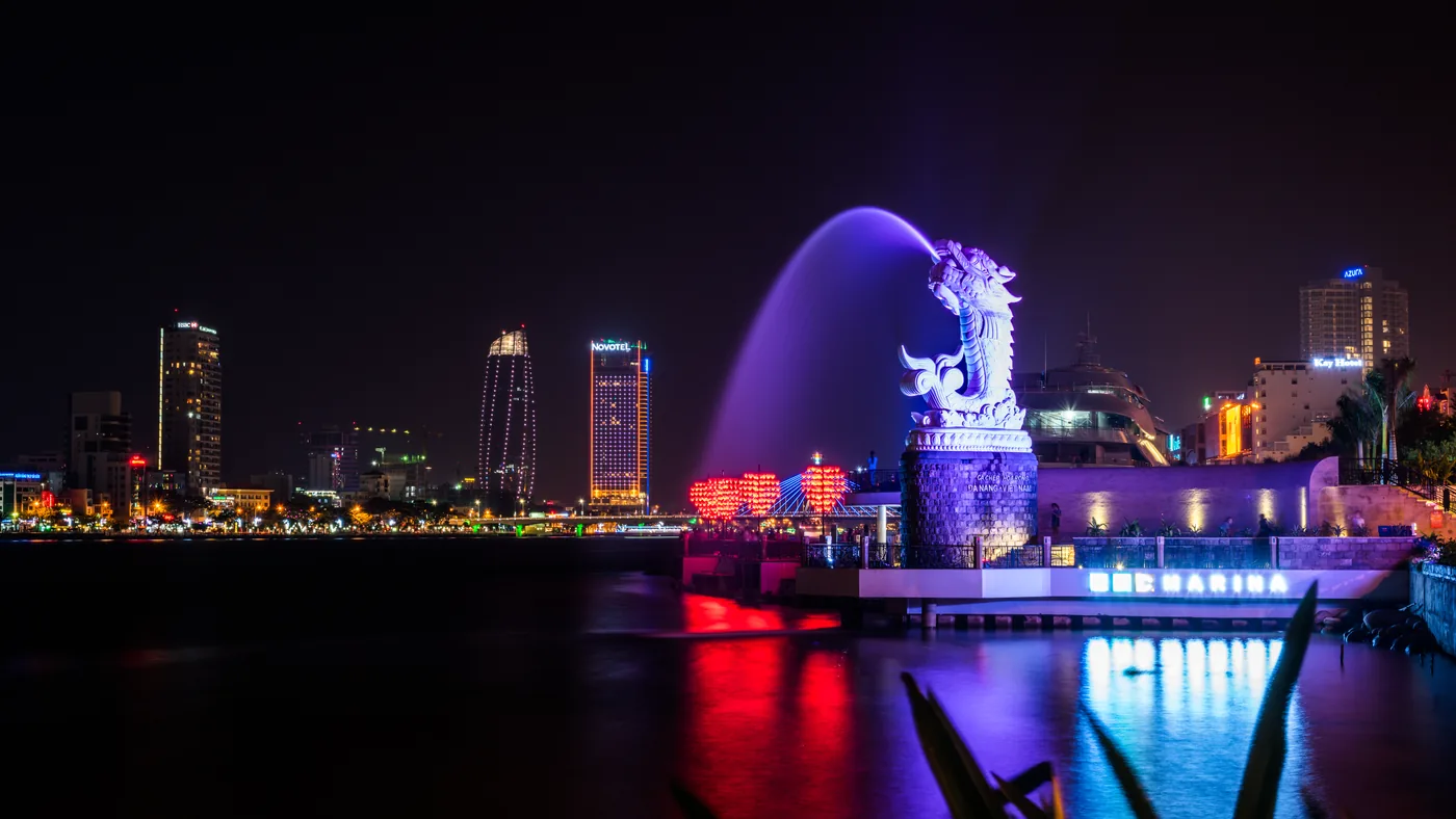 The Carp-to-Dragon statue floodlit purple and spouting a jet of water, with the red heart-shaped arches of the Love Lock Bridge and a DHC Marina sign beside it