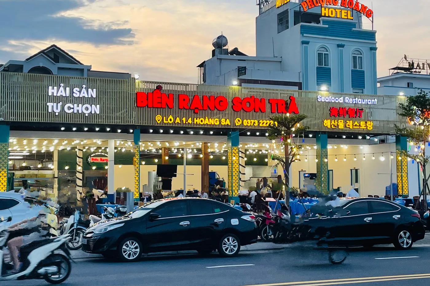 A lit seafood restaurant in Da Nang seen from across the road at dusk, with a large red sign reading BIEN RANG SON TRA and HAI SAN TU CHON, festoon lights and diners inside