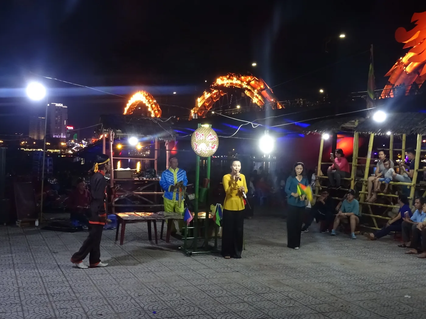 An open-air Bai Choi folk performance on the Da Nang riverfront, with two women singers in ao dai, a man in traditional dress and a crowd of about thirty people, the Dragon Bridge lit gold behind
