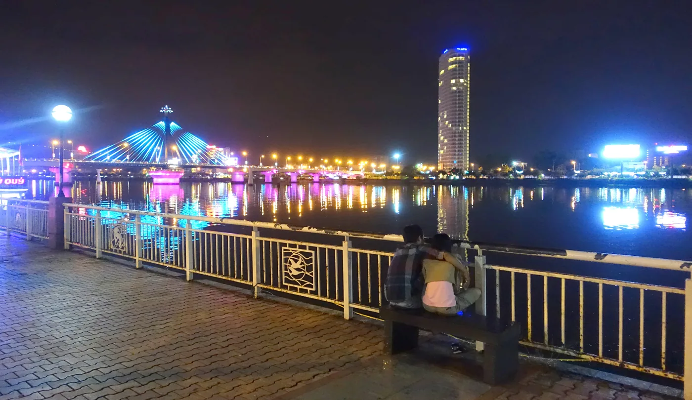 A couple sitting on a stone bench at a railing on the Han River promenade at night, with the Han River Bridge lit blue and pink behind them