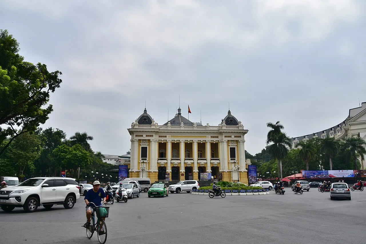 隔著廣場正面拍攝的河內大劇院,黃奶油色的新古典主義立面,前方有車流與一名騎單車的人 隔著廣場正面拍攝的河內大劇院,黃奶油色的新古典主義立面,前方有車流與一名騎單車的人