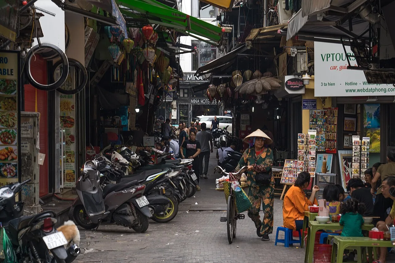 河內老城區靠近陶硯街的窄巷,機車沿著路緣車頭朝內並排,旅行社店面外掛著燈籠與斗笠,一名女子推著腳踏車經過路邊攤的矮板凳 河內老城區靠近陶硯街的窄巷,機車沿著路緣車頭朝內並排,旅行社店面外掛著燈籠與斗笠,一名女子推著腳踏車經過路邊攤的矮板凳