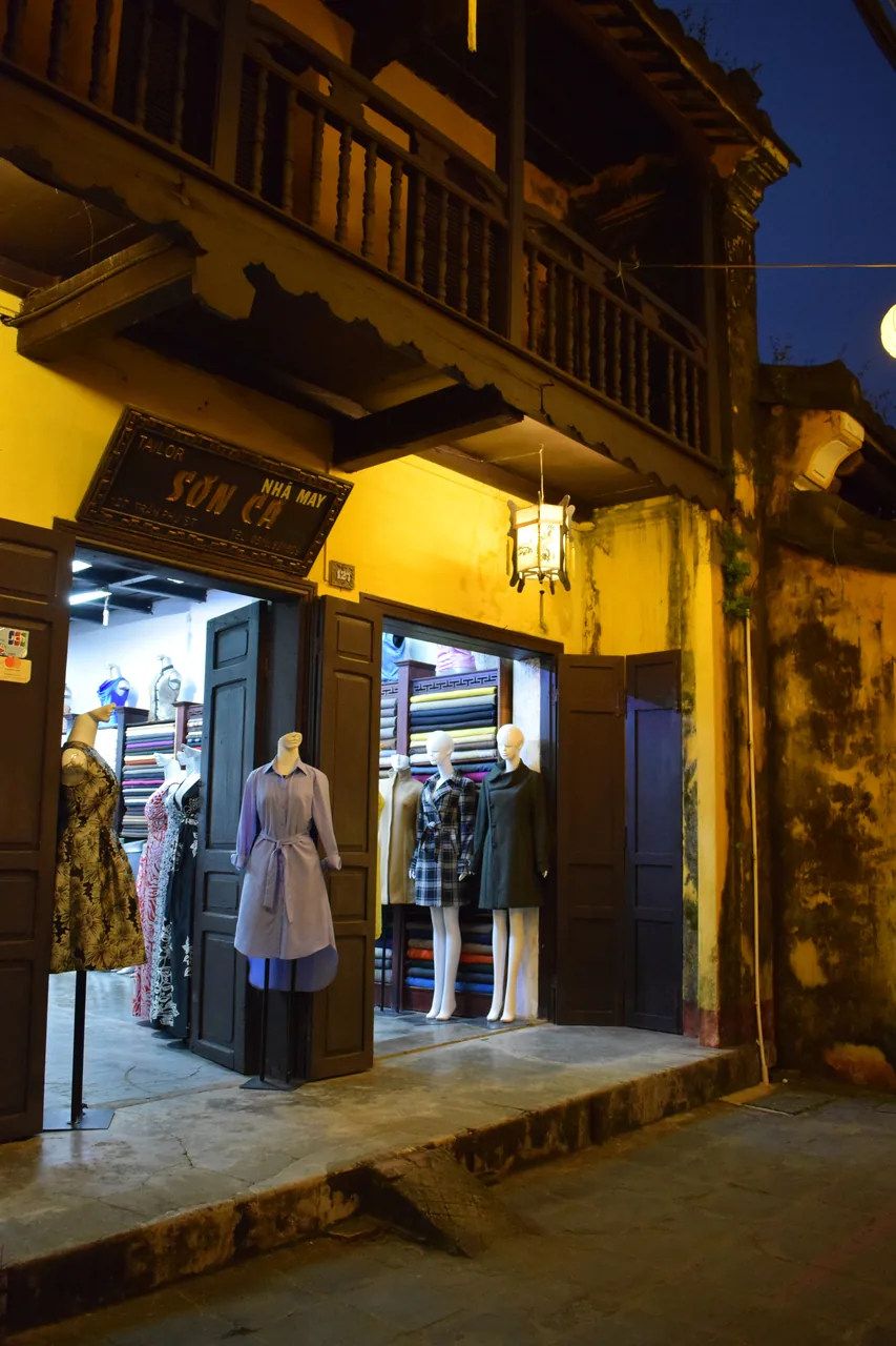 A lantern-lit Hoi An tailor shop at dusk with dresses and bolts of fabric
