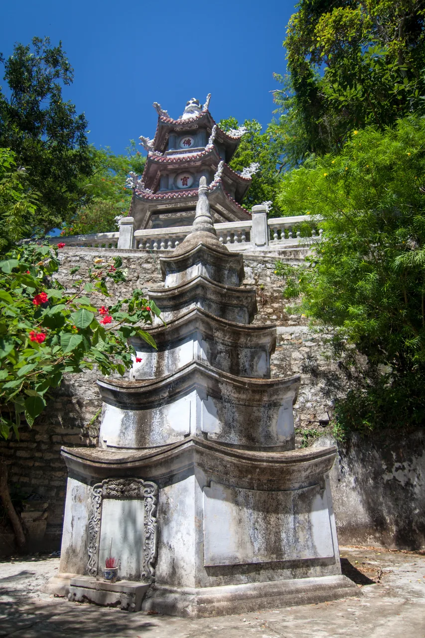 A weathered stone memorial stupa rising in front of a pagoda at the Marble Mountains, framed by red flowers and green trees under a blue sky