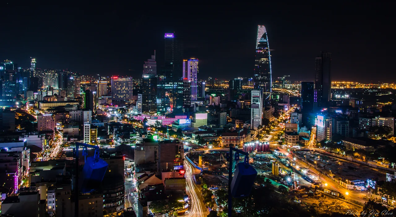 Ho Chi Minh City at night, motorbike light trails streaming past illuminated high-rises and the Bitexco tower.
