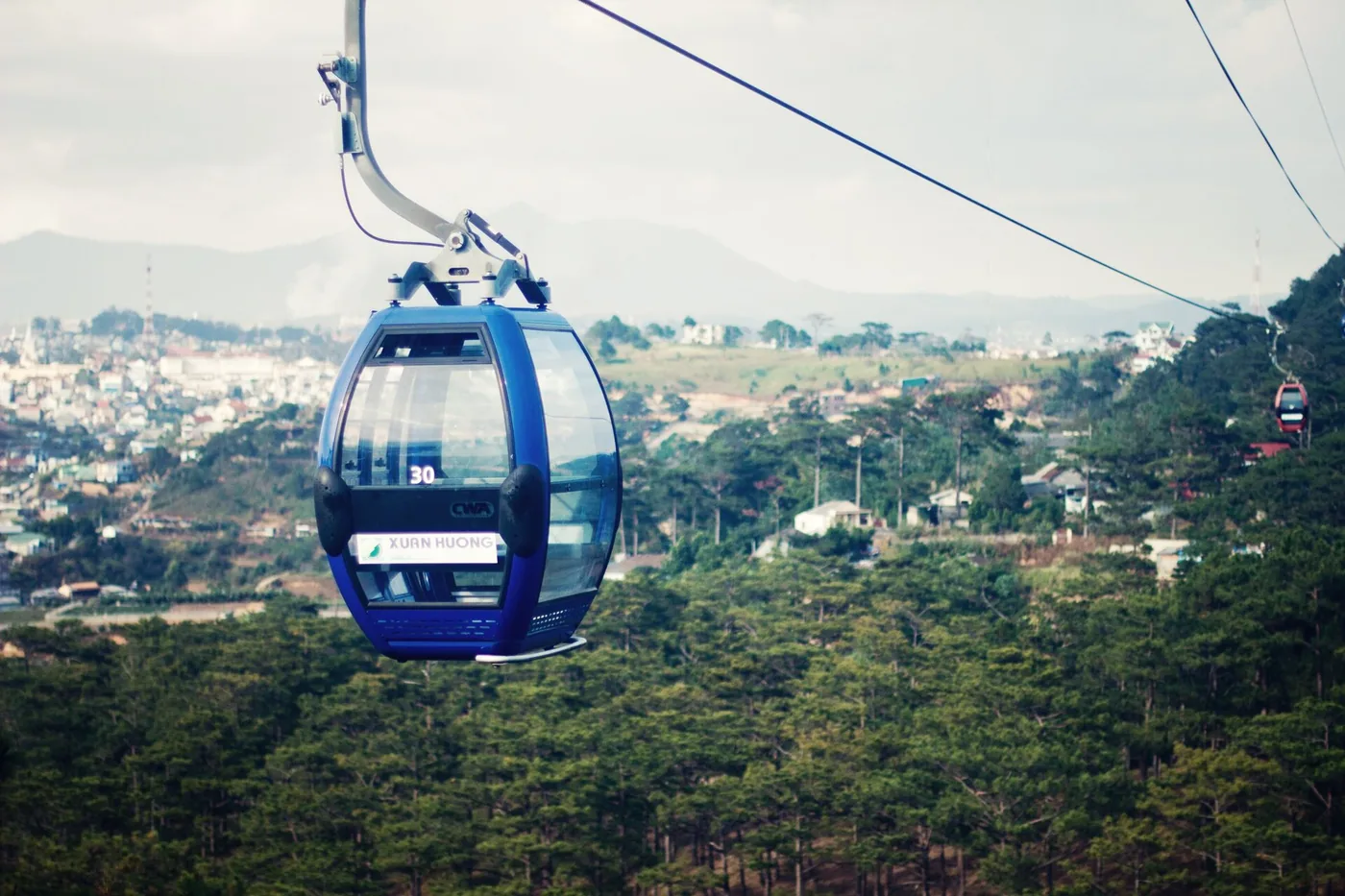 A blue cable car gondola over pine forest with Da Lat in the valley below