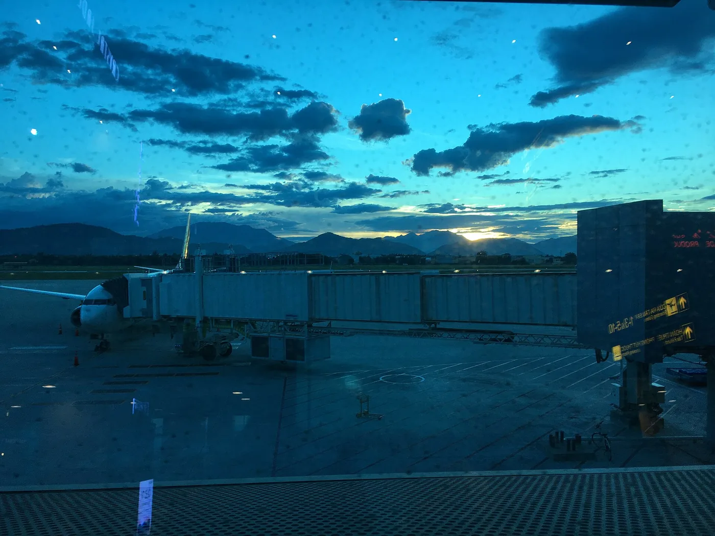 The apron at Da Nang international airport after sunset, seen through a terminal window with an aircraft at the jet bridge and the mountains behind