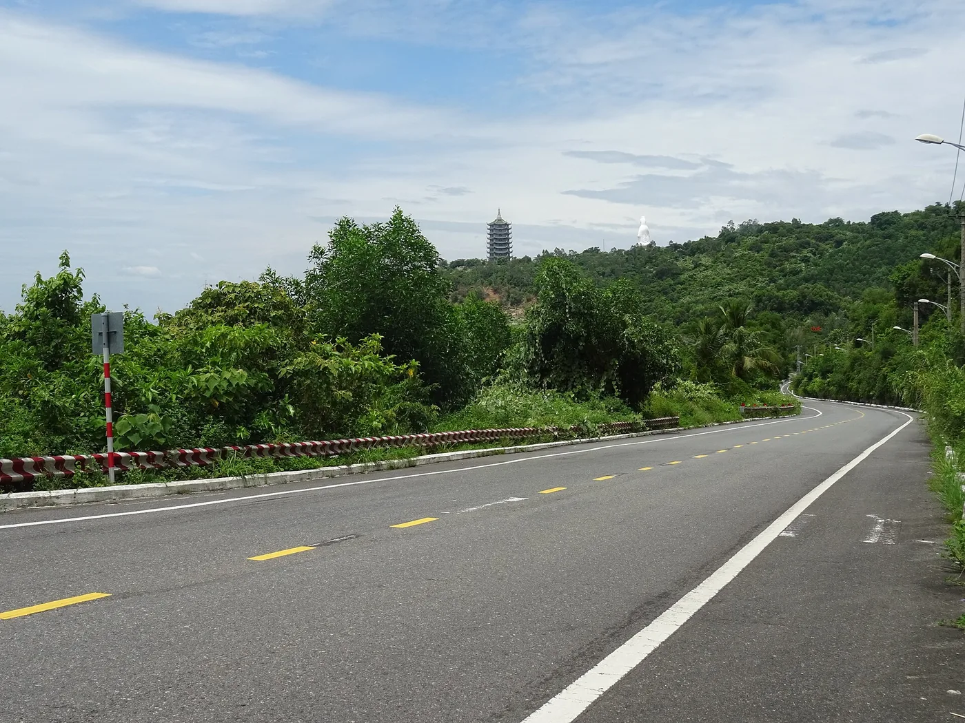 An empty road curving up the Son Tra peninsula near Da Nang, with a pagoda tower and a white statue visible on the wooded ridge above