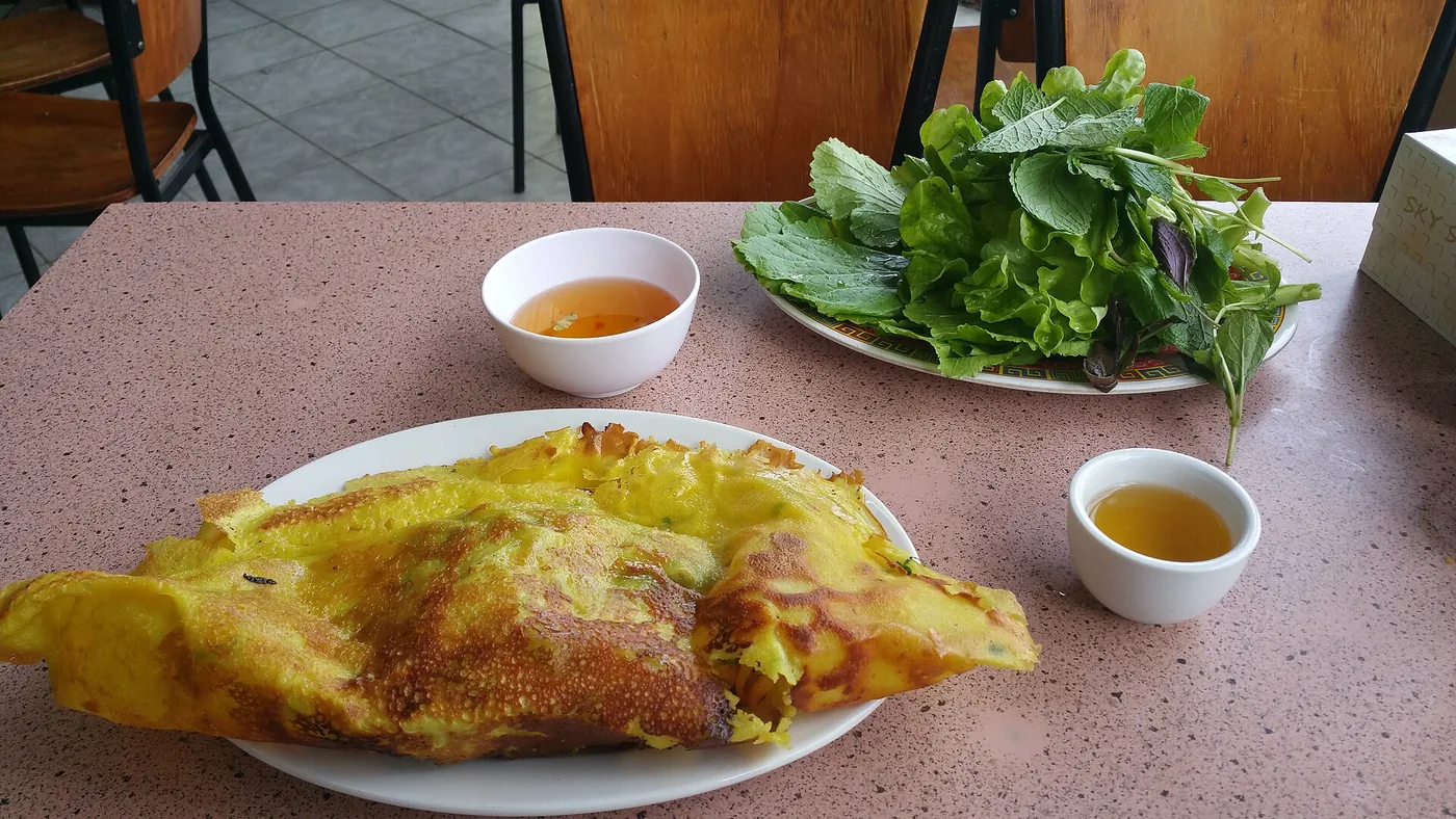 One banh xeo pancake on a plate next to a plate of lettuce and herbs and two small bowls of dipping sauce