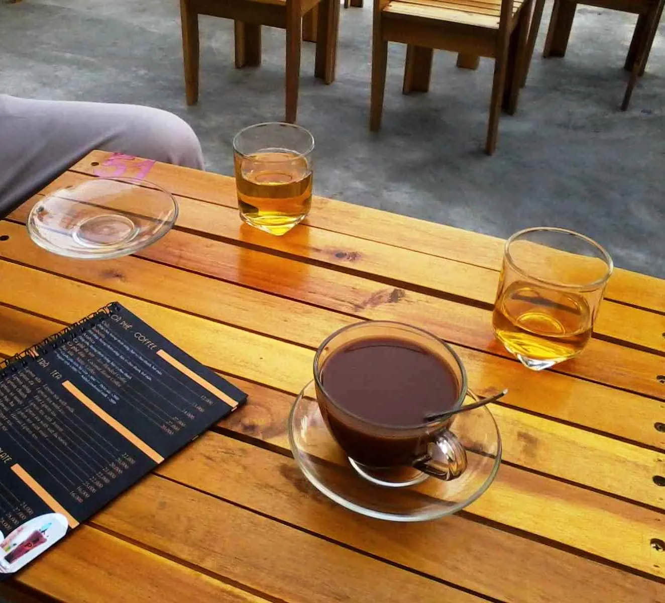 A wooden cafe table in Da Nang with one glass cup of black coffee, two glasses of iced tea and a printed drinks menu