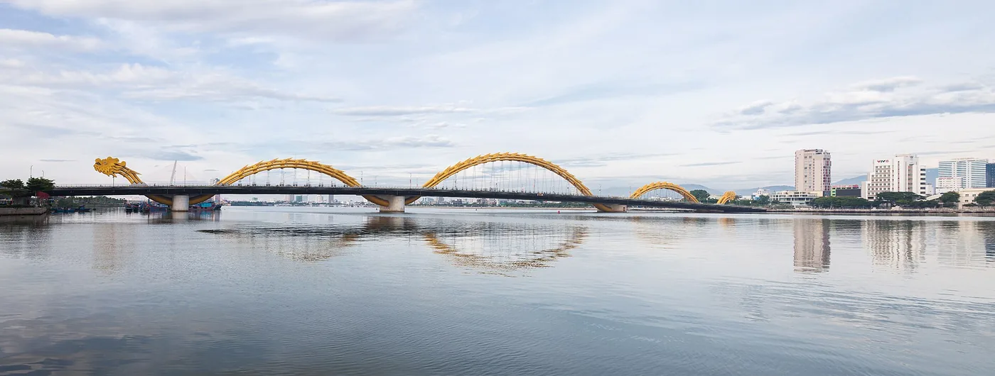 The Dragon Bridge in Da Nang in daylight, its yellow dragon frame crossing the Han River and reflected in the water