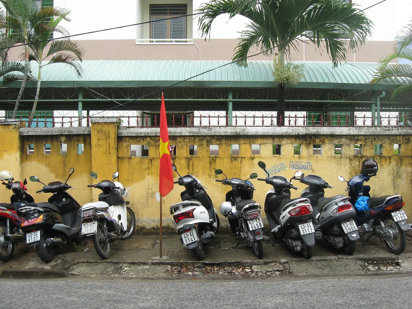 A row of scooters parked along a yellow wall on a Da Nang street with a Vietnamese flag between them