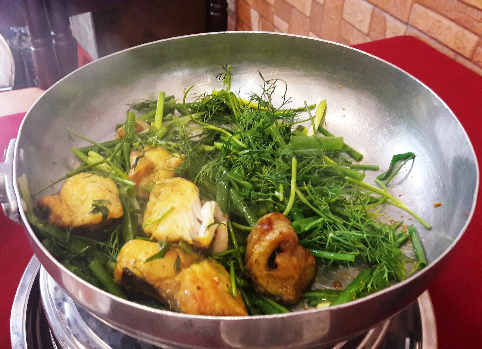 Turmeric-marinated fish cooking in a pan at the table with dill and spring onion piled on top