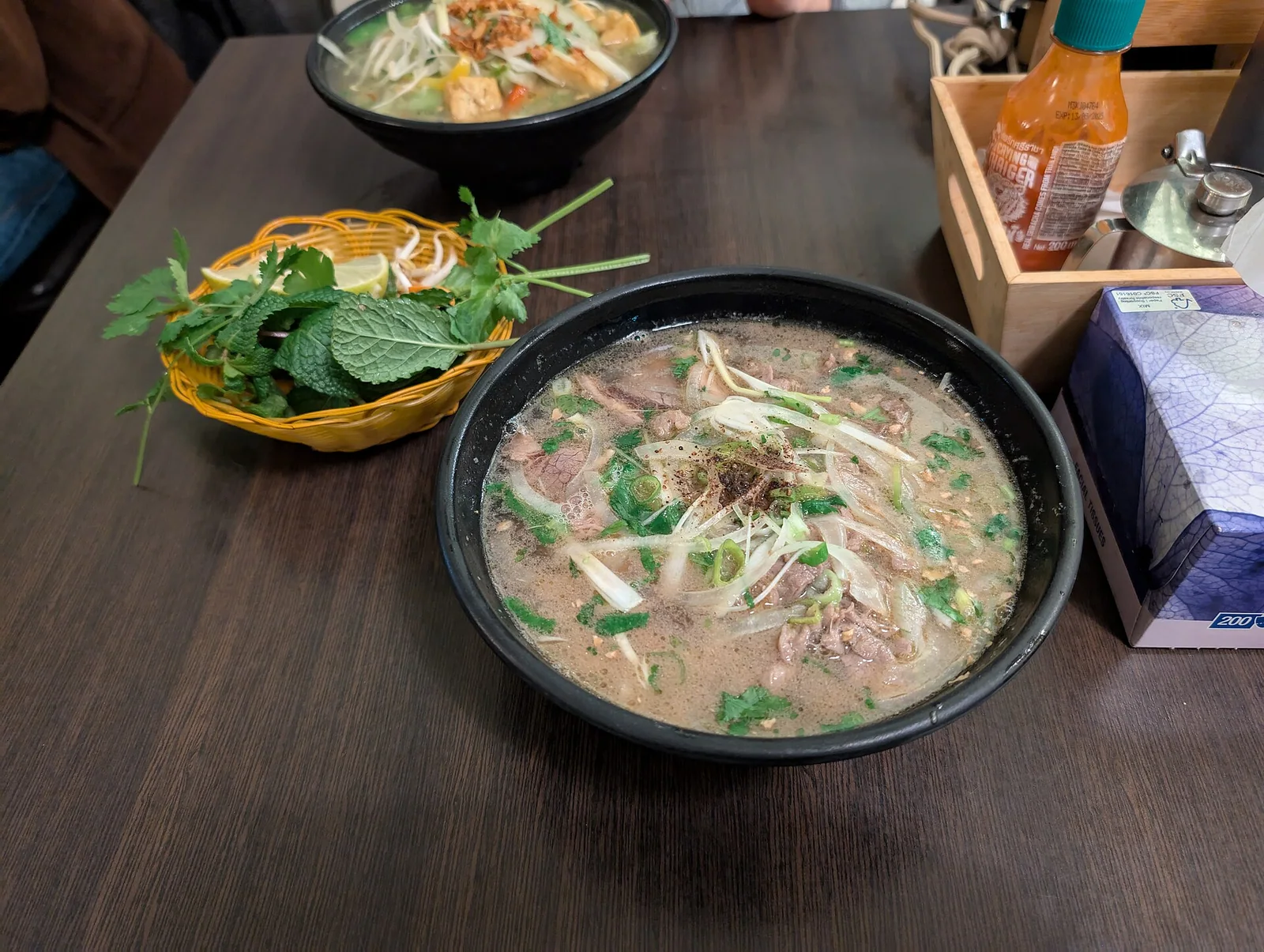 A bowl of Hanoi beef pho with clear broth, flat rice noodles and fresh herbs on the side