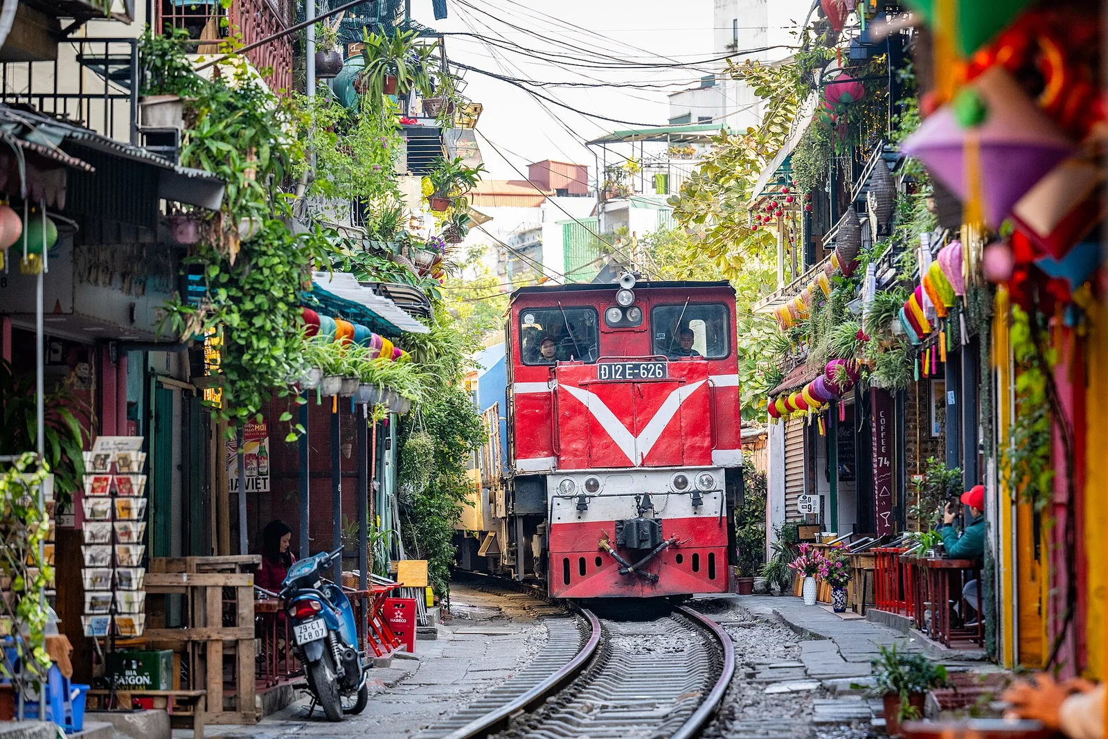 A red train passing along a narrow railway line between houses on the train street in Hanoi