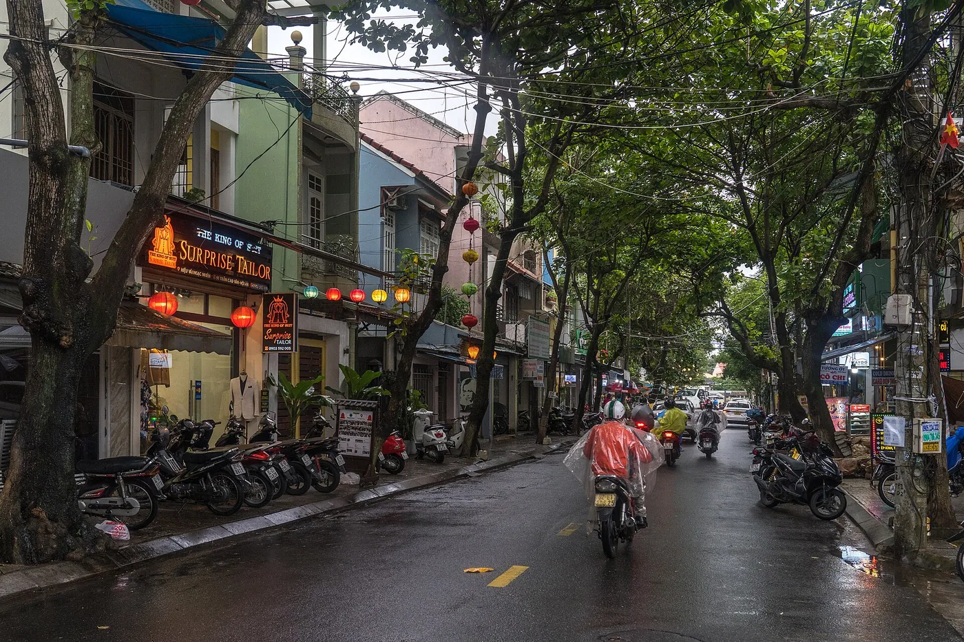 A wet street in Hoi An in November, riders in rain ponchos passing parked scooters with lanterns strung between the trees