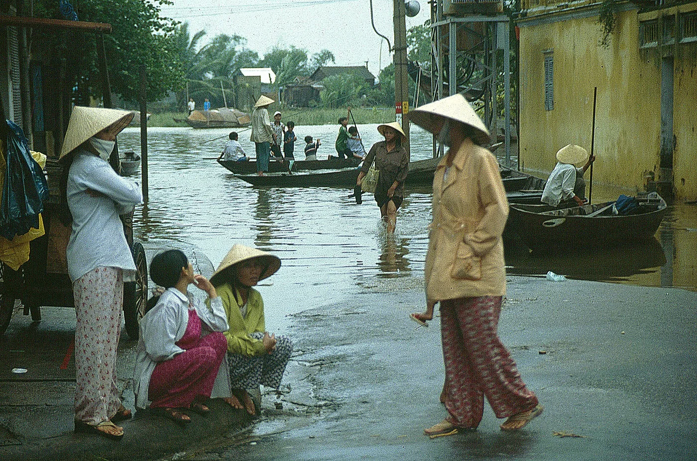 A flooded street in Hoi An with wooden boats being poled along it while people in conical hats stand on the dry end of the pavement