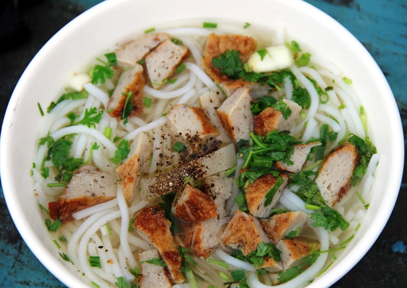 A white bowl of Nha Trang bun cha ca: thick white rice noodles in a clear broth with slices of steamed and fried fish cake, coriander and spring onion on top