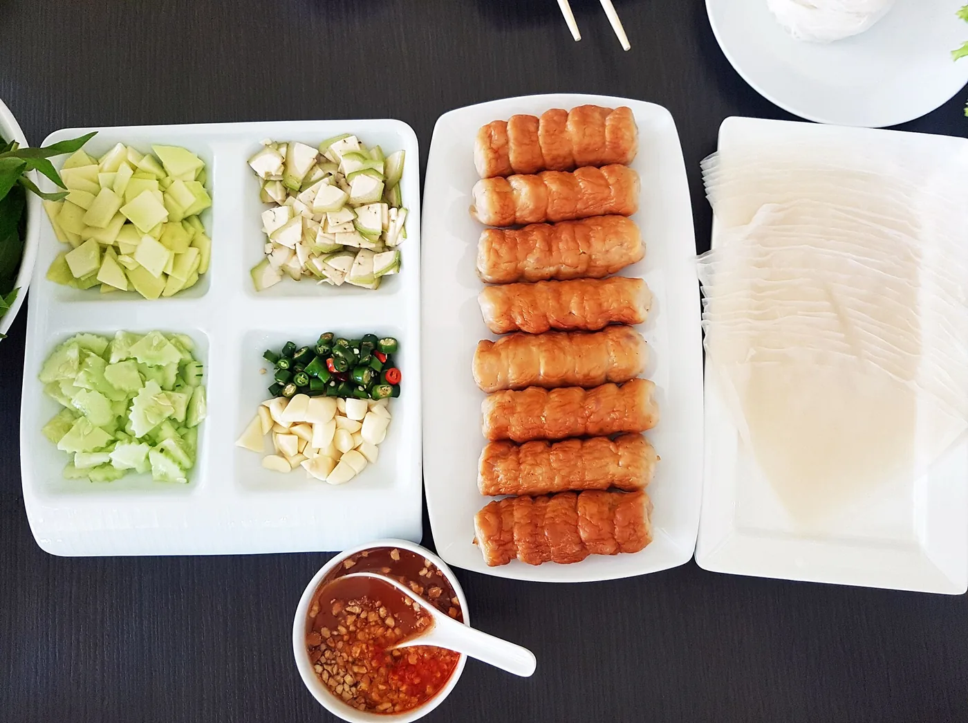 흰 접시에 차려진 넴느엉 한 상, 구운 돼지고기 롤이 줄지어 있고 라이스페이퍼와 파란 망고·오이·고추·마늘, 걸쭉한 소스 그릇이 함께 놓여 있다