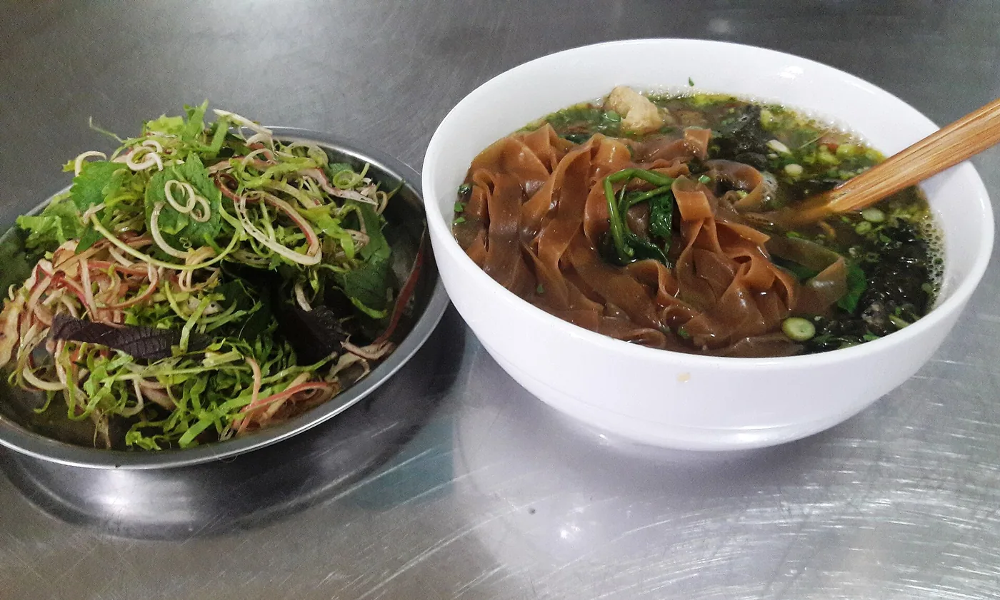 A bowl of banh da cua next to a plate of fresh herbs
