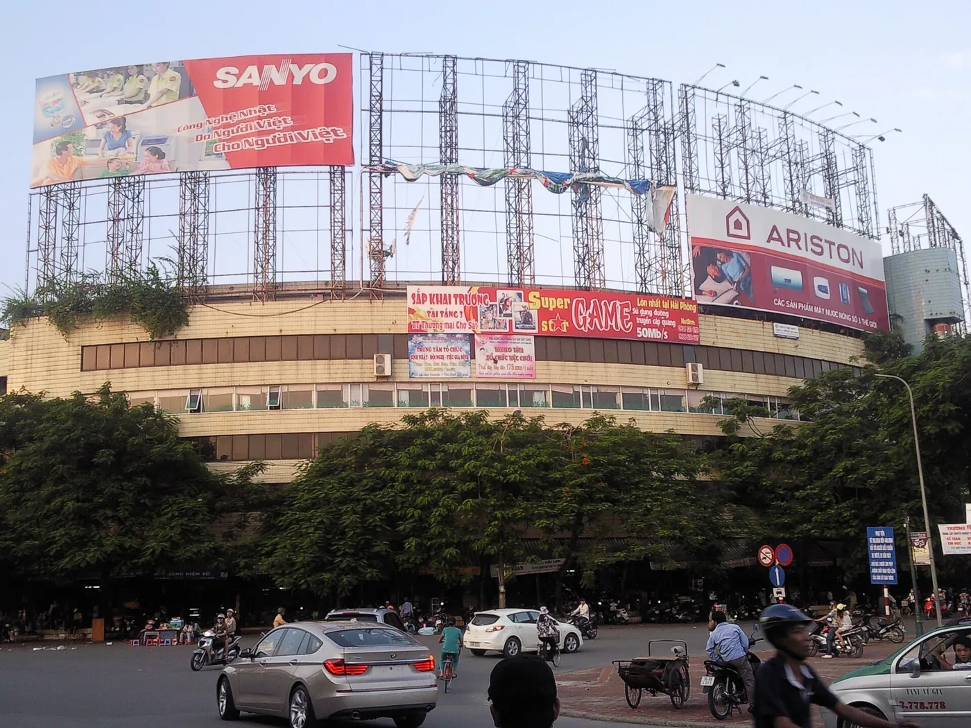 The front of Cho Sat market, an ageing block hung with large signs, with cars and motorbikes passing