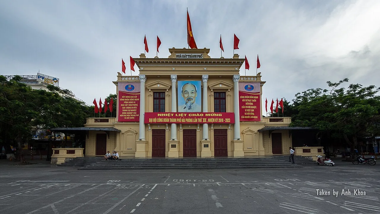 The front of Hai Phong Opera House, a yellow building hung with red flags, facing an empty square