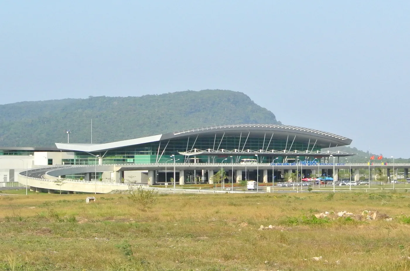 The terminal at Phu Quoc International Airport, a long curved roof with a green hill behind it and a raised approach road in front